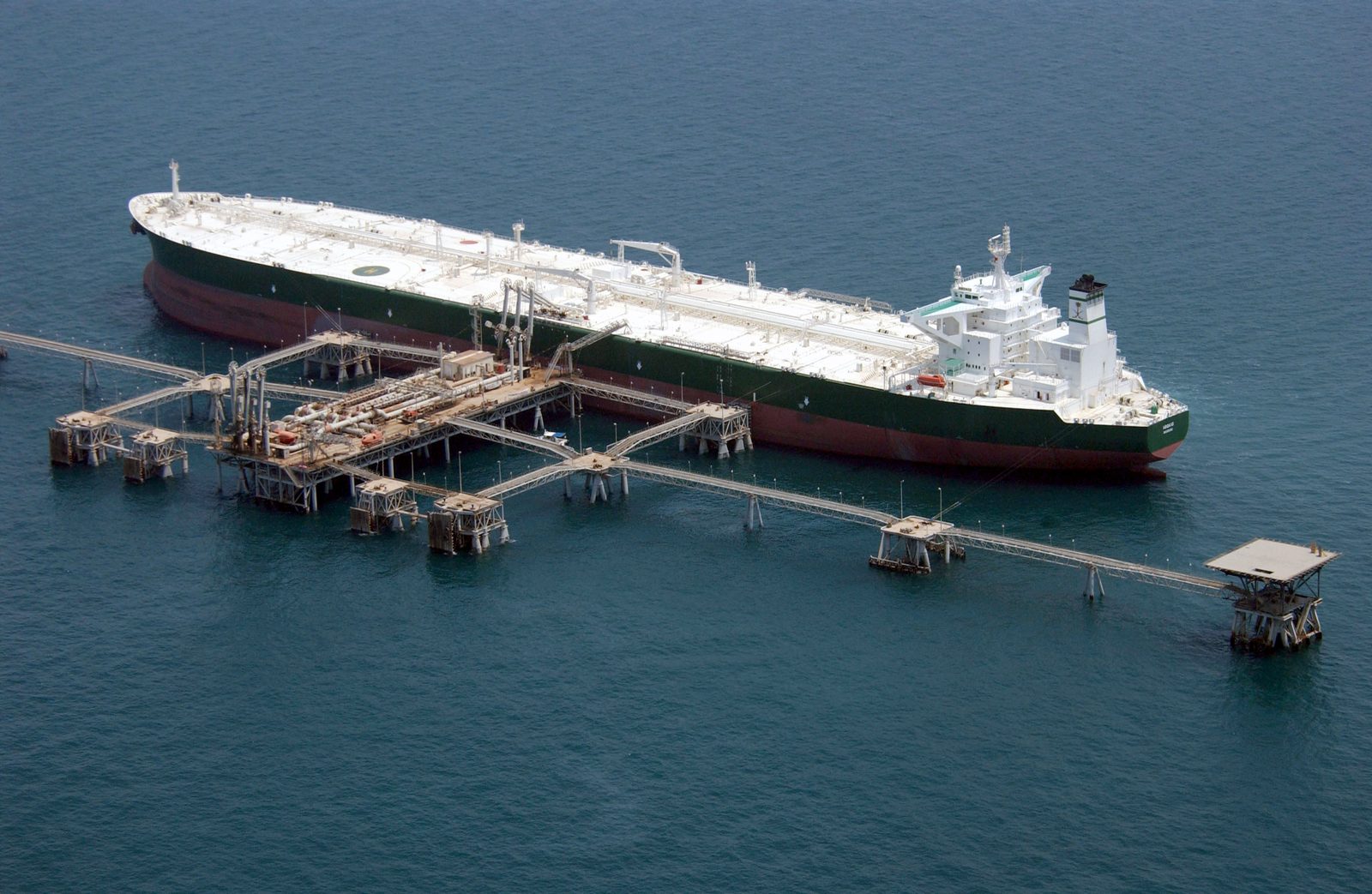 A supertanker moored at an offshore oil terminal in the Persian Gulf, seen from above, with loading arms and walkways connecting it to the platform