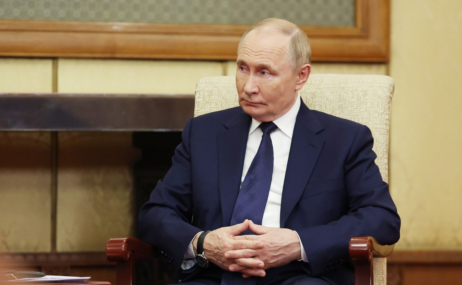 Vladimir Putin, seated in a beige armchair in a dark suit and blue tie, hands clasped, during a meeting in September 2025